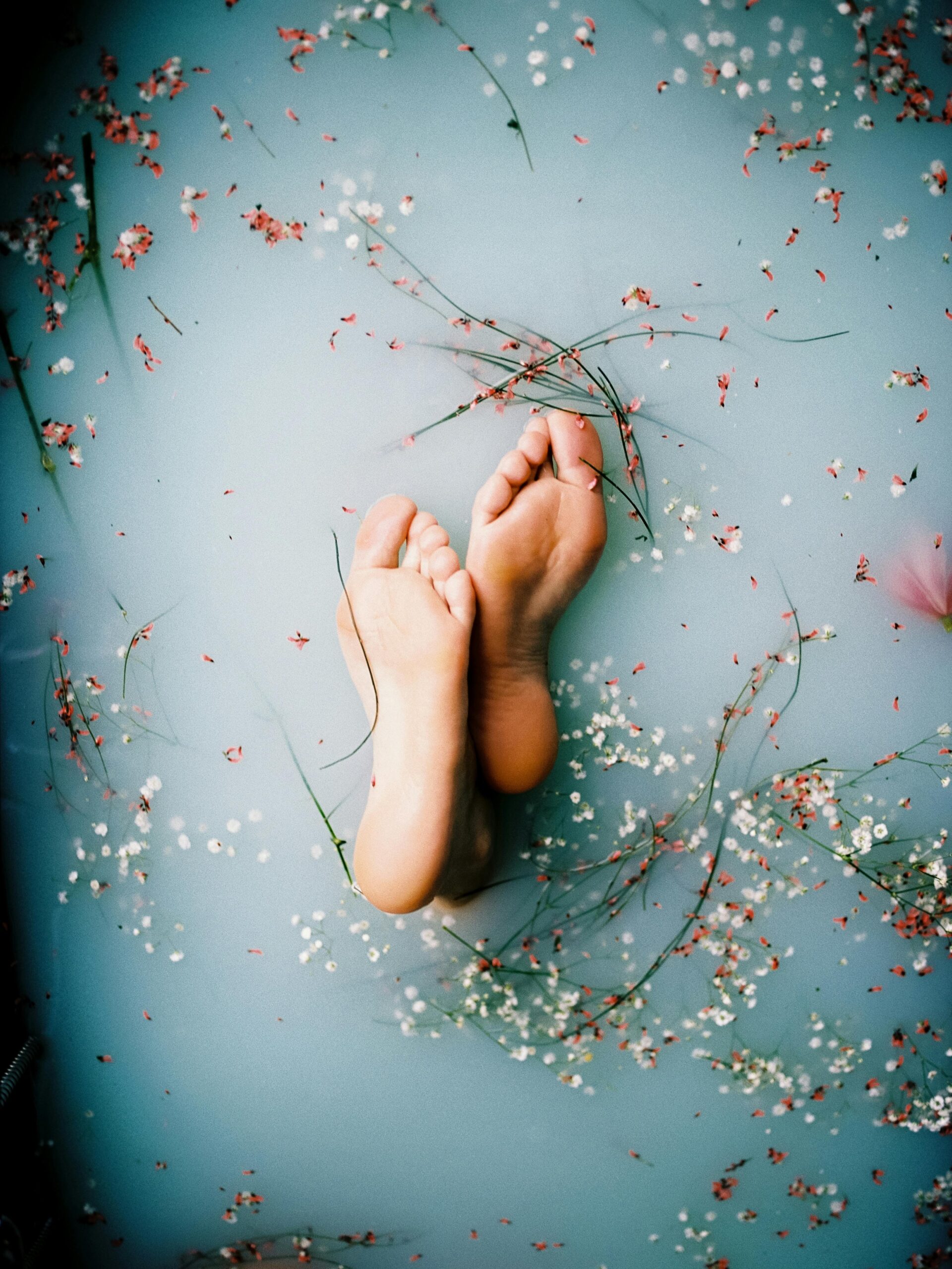 Bare feet submerged in pastel water adorned with delicate petals and flowers, creating a tranquil scene.