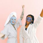 Happy diverse women in robes and blue collagen masks applied on face standing with raising hands near pale pink wall and looking at camera