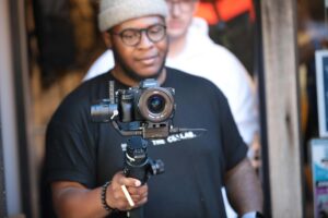 Black male videographer with a modern camera filming indoors, capturing a laid-back scene.